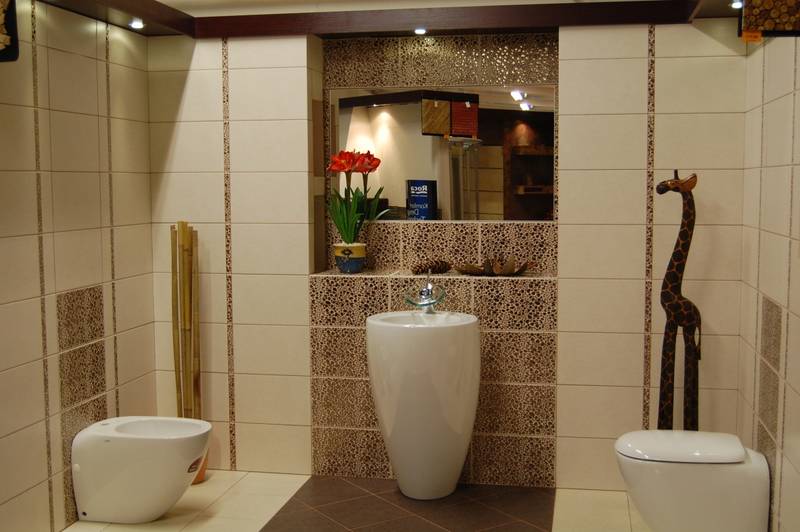 Jasna łazienka z beżowymi i brązowymi płytkami, wolnostojącą umywalką, bidetem, toaletą i drewnianą figurką żyrafy jako elementem dekoracyjnym.